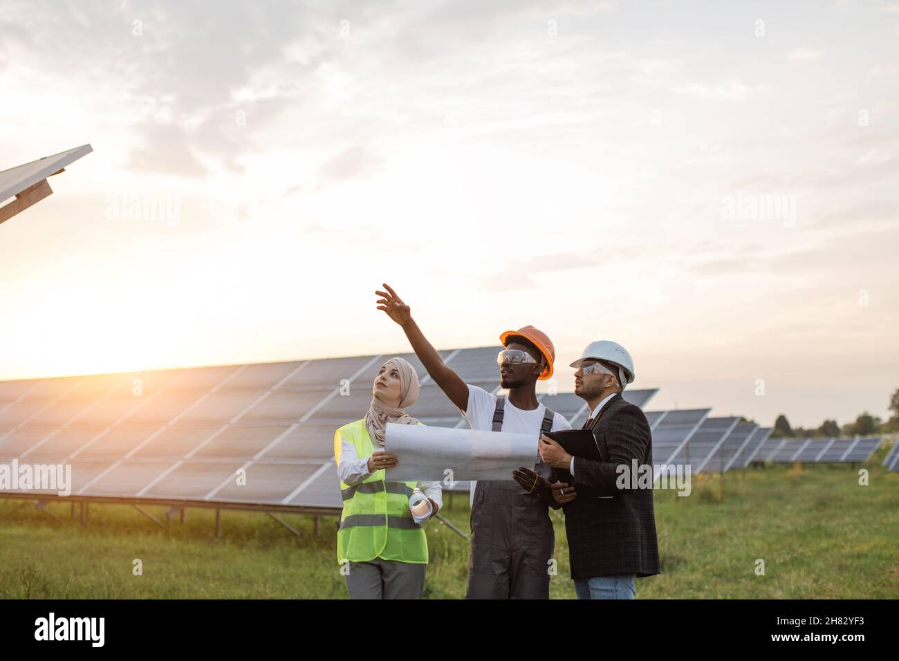 afroamerikanische Technikerin im Gespräch mit muslimischer Frau und indischem Mann während der Arbeitssitzung auf einer Solarstation. Industriearbeiter diskutieren gemeinsames Projekt der grünen Energieerzeugung. Stockfoto