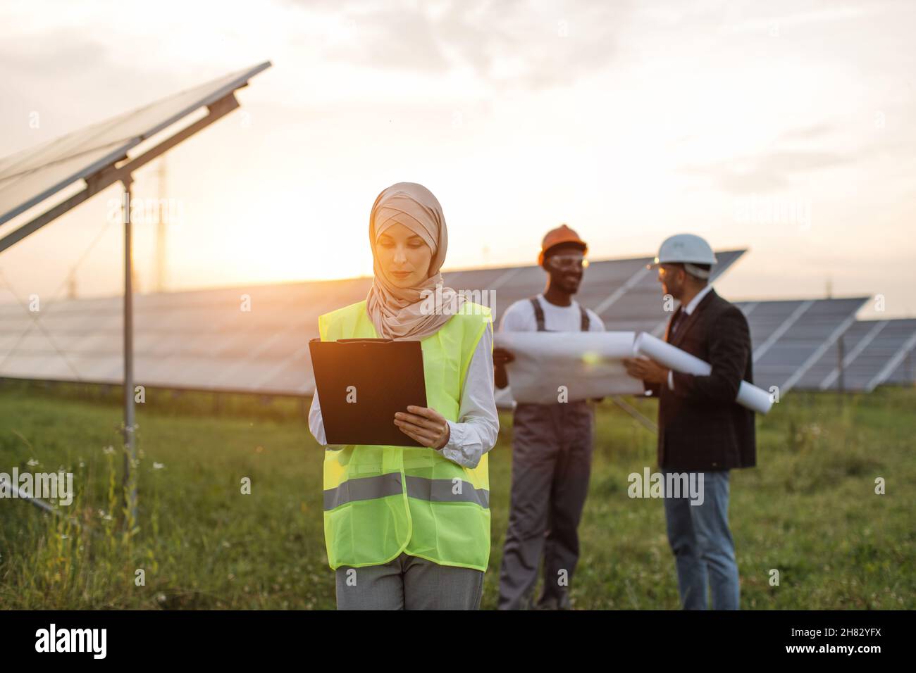 Die muslimische Frau hijab schrieb auf die Zwischenablage, während ihre männlichen Kollegen hinter Helmen standen und Blaupausen untersuchten. Multirassische Menschen, die die Produktion von Sonnenenergie entwickeln. Stockfoto