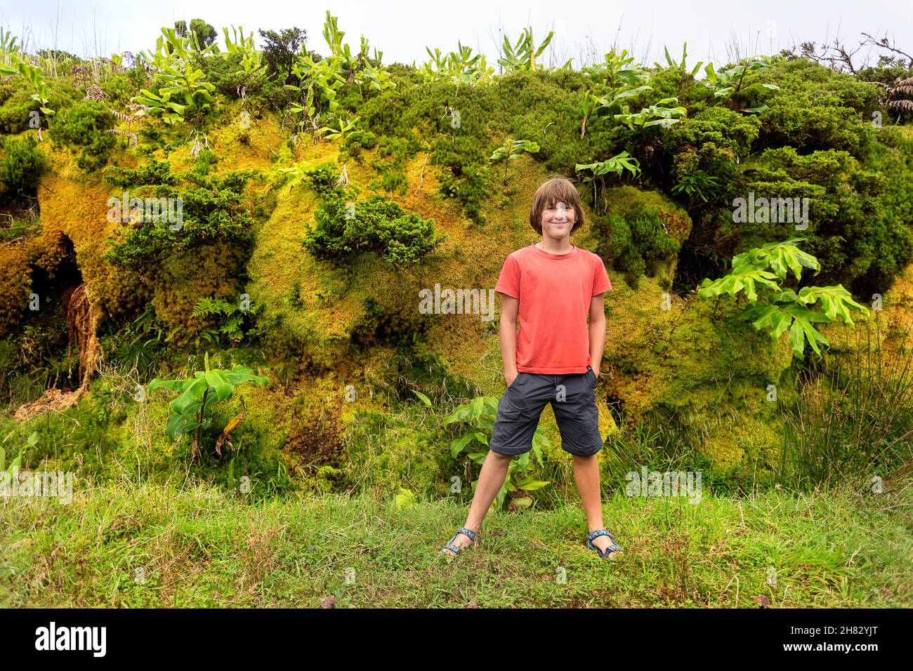 Lächelnder Junge mit Händen in Taschen, der vor farnbedeckten Wänden steht, Flores, Azoren, Portugal Stockfoto