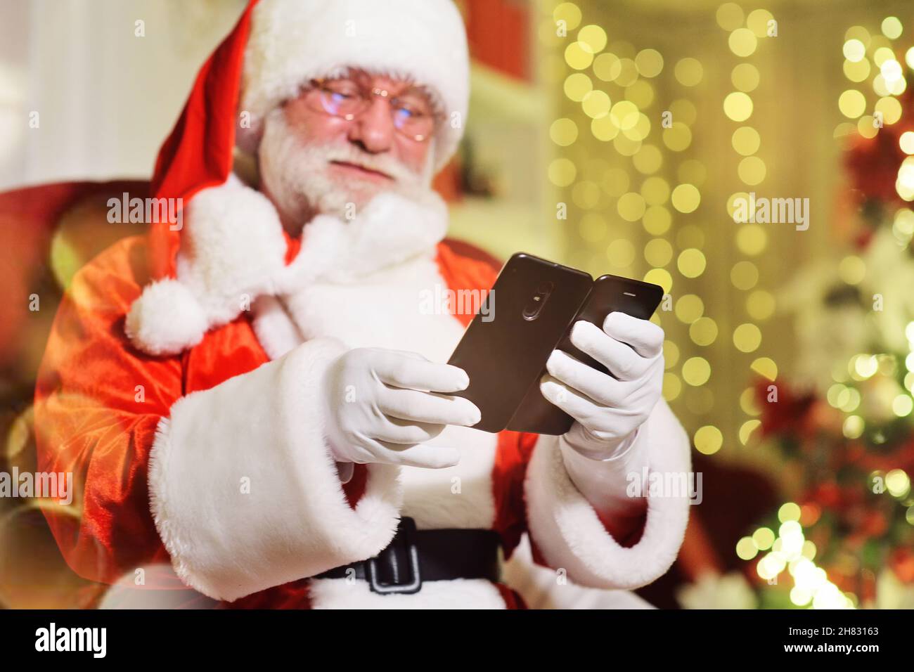 Der Weihnachtsmann mit einem echten grauen Bart in einem traditionellen Kostüm sitzt auf einem Stuhl und hält ein Smartphone in den Händen. Der Weihnachtsmann auf der Fernbedienung gratuliert Stockfoto