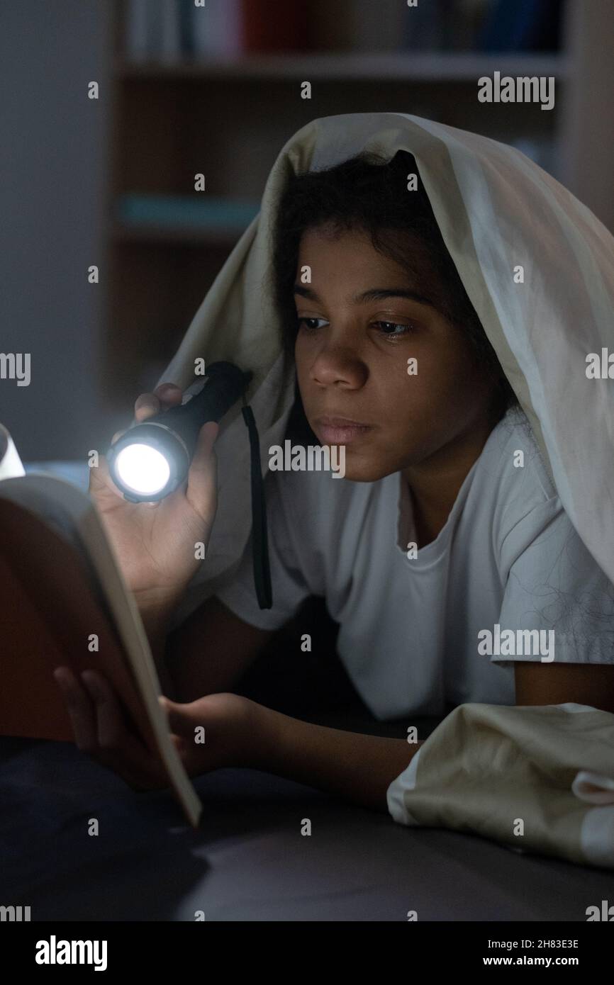 Konzentriertes schwarzes Mädchen unter Bettdecke liegen und mit Taschenlampe beim Lesen Märchen in der Dunkelheit Stockfoto