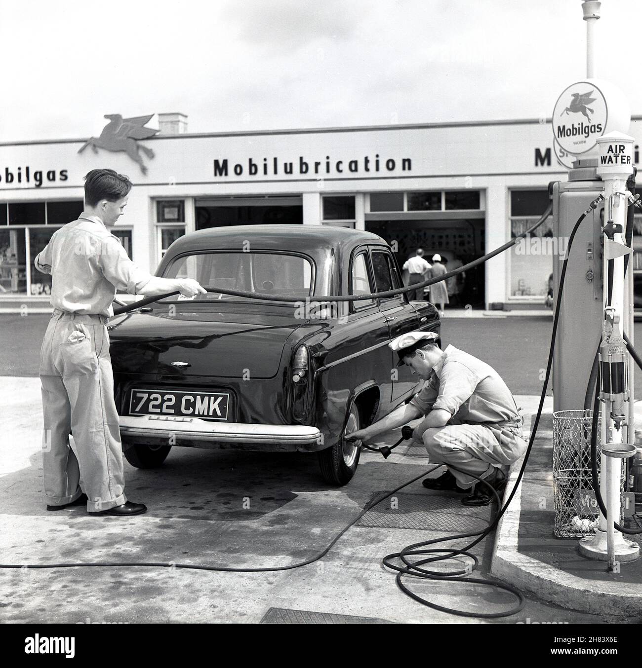 1950s, zwei uniformierte Vorhofmitarbeiter an einer neuen Mobilgas-Tankstelle, neben einem Auto in Austin, das von einem mit Benzin befüllt und der andere seinen Reifendruck überprüft, England, Großbritannien. Ein amerikanisches Ölunternehmen, Vacuum Oil, hat 189 die Marke „Mobilgas“ eingetragen. Sie fusionierte 1931 mit Socony und wurde 1955 zur Socony Mobil Oil Company. Die ersten britischen Tankstellen wurden Anfang 1950s eröffnet und waren voll mit Pumpenwartern und Mechanikern besetzt. In den Nachkriegsjahren stieg der Kfz-Absatz deutlich an, und Großbritannien wurde nach den USA zum zweitgrößten Automobilhersteller der Welt. Stockfoto