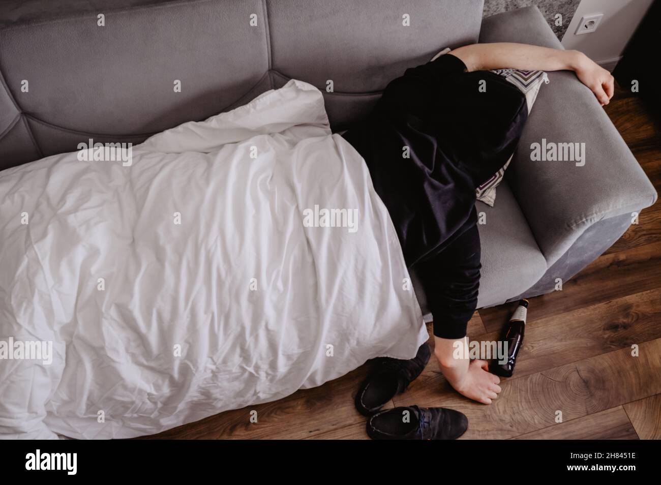 Mann, der mit Kopfschmerzen oder einem Kater auf einer Couch liegt, mit einer Flasche Bier und einer weißen Decke Stockfoto
