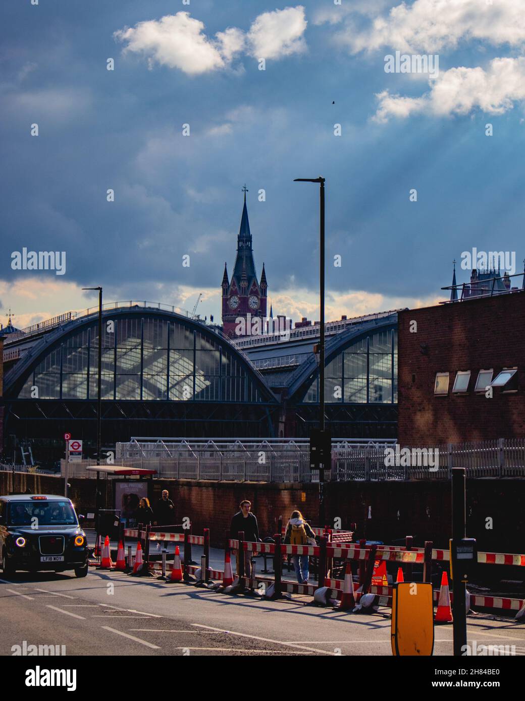 Aufnahme von Kings Cross Station, London, von hinten im Sommer unter Sommerhimmel mit Lichtstrahlen von der Sonne, während ein schwarzes Taxi vorbei fährt und die Leute gehen. Stockfoto