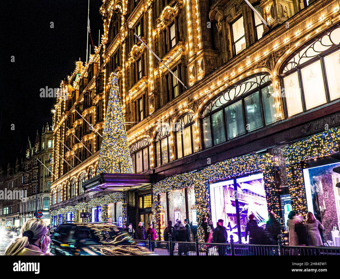 HARRODS CHRISTMAS SHOPPING Einkaufen Londoner Kaufhaus Harrods in der Dämmerung mit Weihnachtsbeleuchtung Menschenmengen und vorbeifahrende Autos Knightsbridge London SW1 Stockfoto