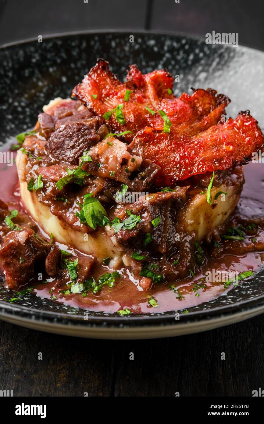 Nahaufnahme von Rindergulasch mit Speckchips, Kartoffelpüree und schwarzer Johannisbeersoße auf einem Teller Stockfoto