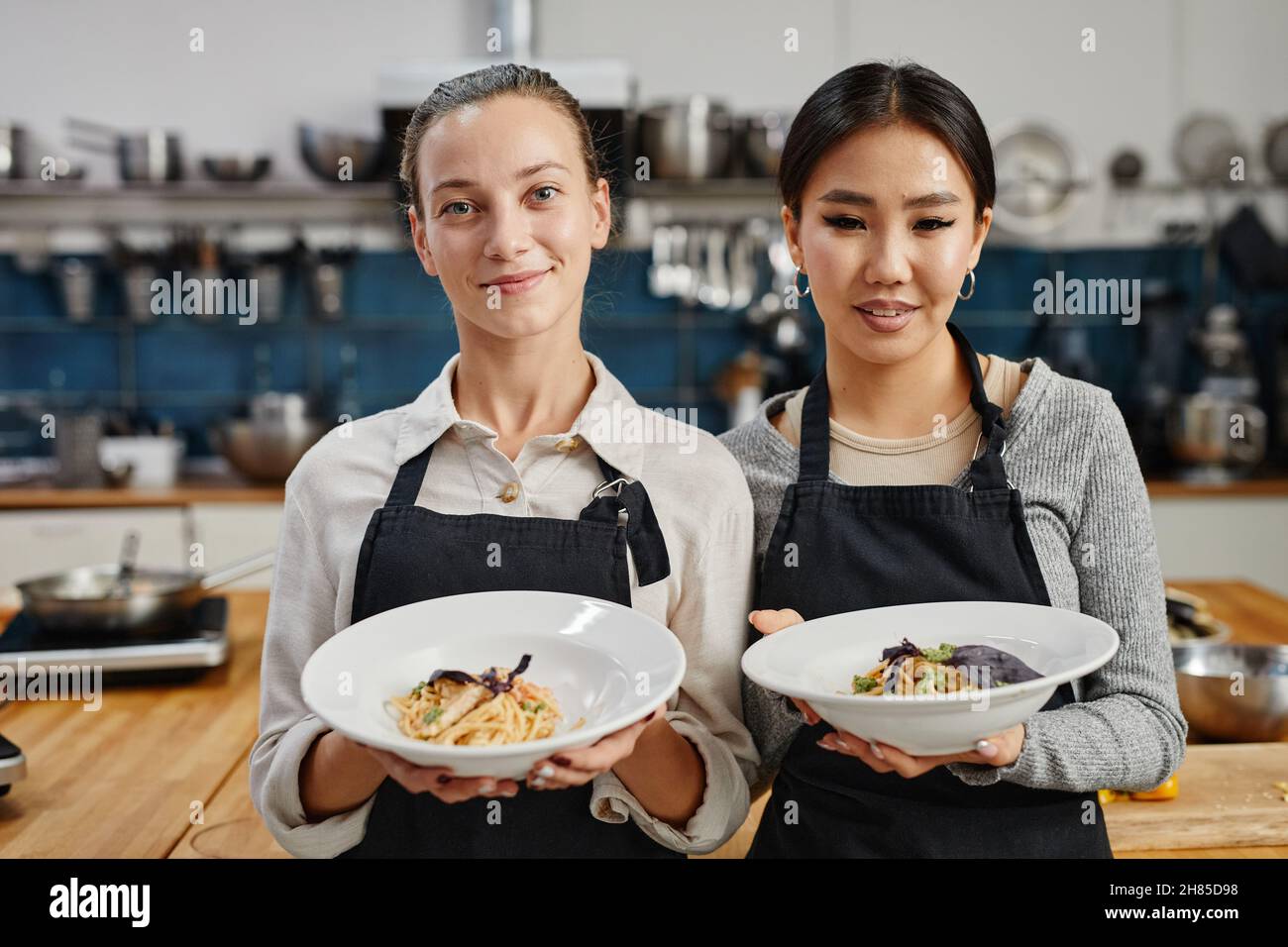 Porträt von zwei lächelnden jungen Frauen, die Schürzen tragen und Gourmet-Gerichte in der professionellen Küche halten, Kopierraum Stockfoto