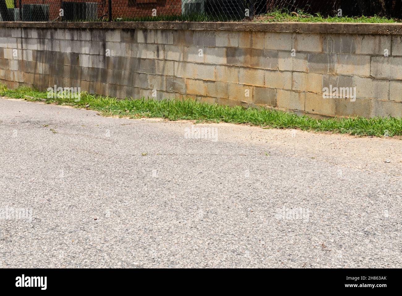 Betonblock Stützmauer entlang einer asphaltierten Auffahrt, gekrönt mit Gras und Kettengliederzaun, horizontaler Aspekt Stockfoto