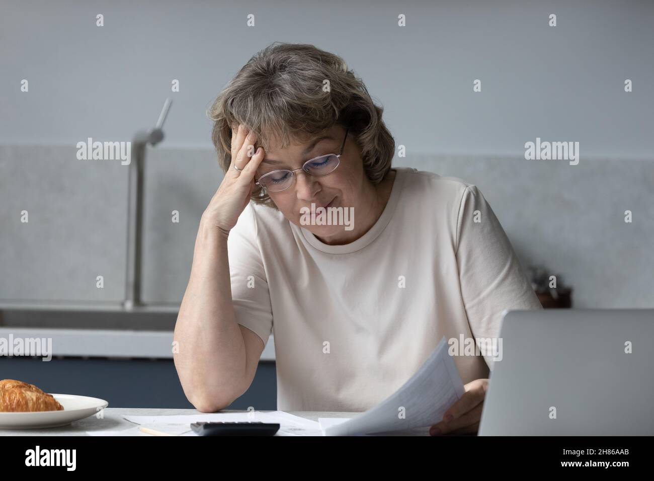 Besorgte Hausbesitzerin mittleren Alters, die am Laptop Papier liest Stockfoto