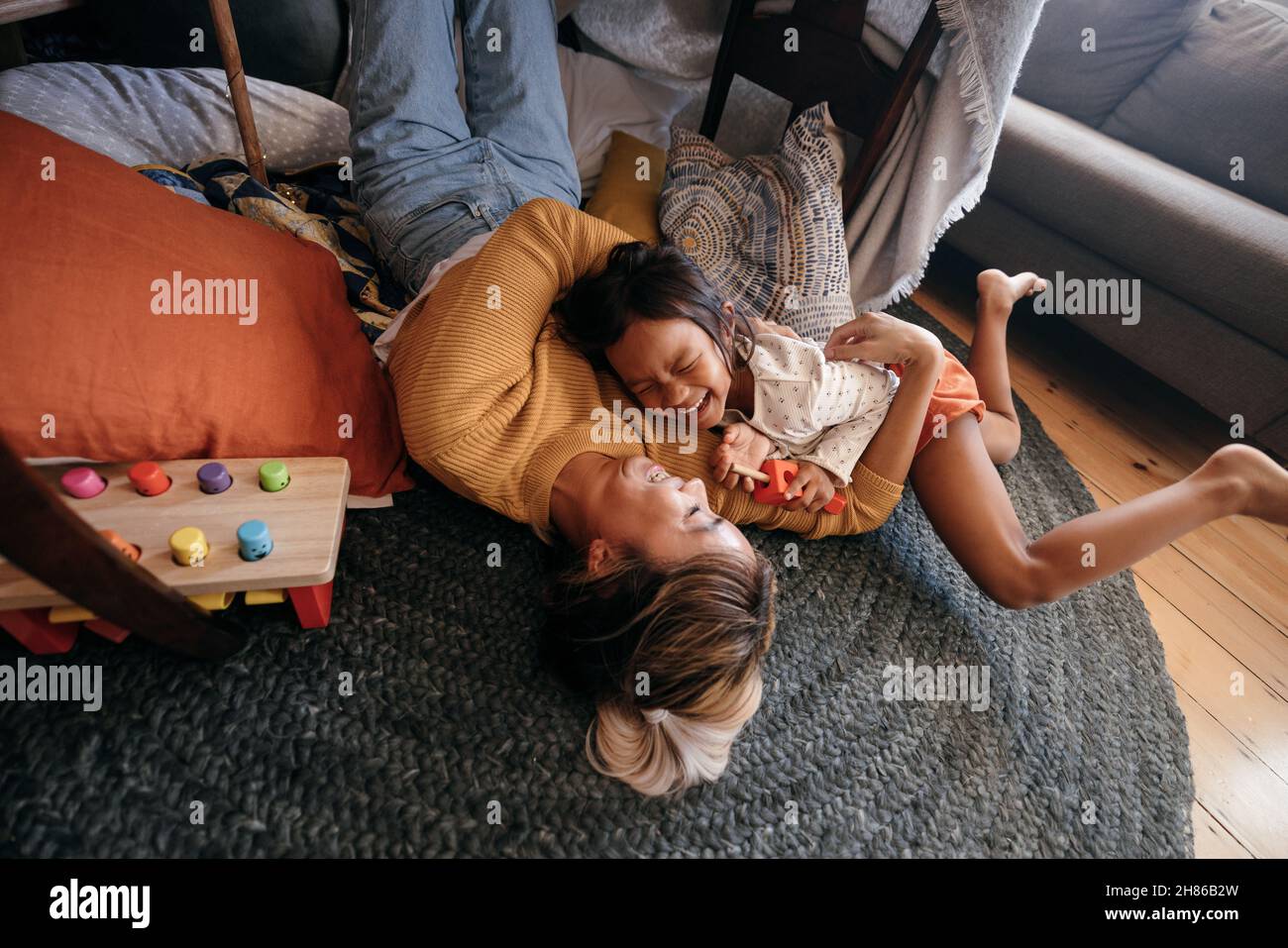 Blick aus der Ferne auf eine Mutter und ihre Tochter, die zu Hause fröhlich lachen. Fröhliche Mutter und Tochter liegen auf dem Boden in ihrem Spielbereich. Mutter und Stockfoto