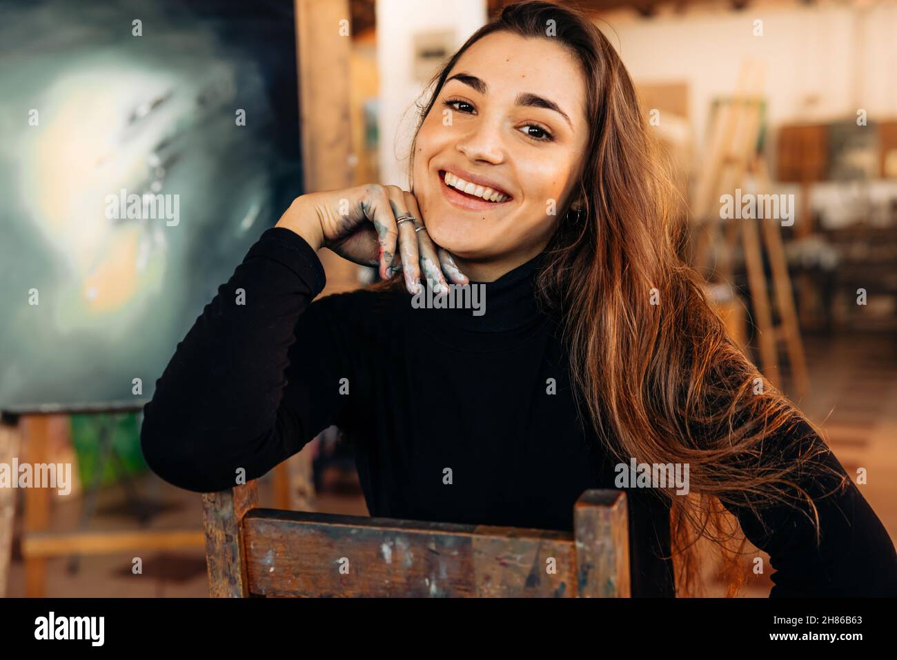 Glückliche professionelle Malerin lächelt der Kamera in ihrem Arbeitsateliersvor. Erfolgreiche Künstlerin sitzt auf einem Stuhl in ihrer Werkstatt mit ihrem Kunstwerk im b Stockfoto