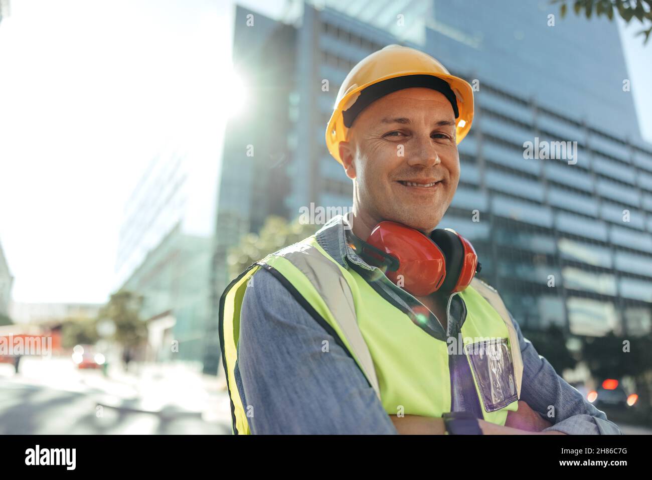 Glücklicher Bauarbeiter, der die Kamera lächelt, während er mit gekreuzten Armen in der Stadt steht. Arbeiter mit blauem Kragen, der vor Hi steht Stockfoto