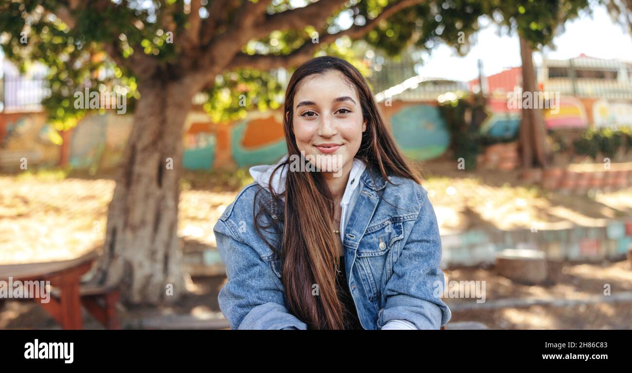 Selbstbewusstes Teenager-Mädchen, das mit einem Lächeln auf die Kamera schaut. Fröhliche Brünette Mädchen trägt eine Denim-Jacke im Freien in der Stadt. Weibliche junge Sittin Stockfoto