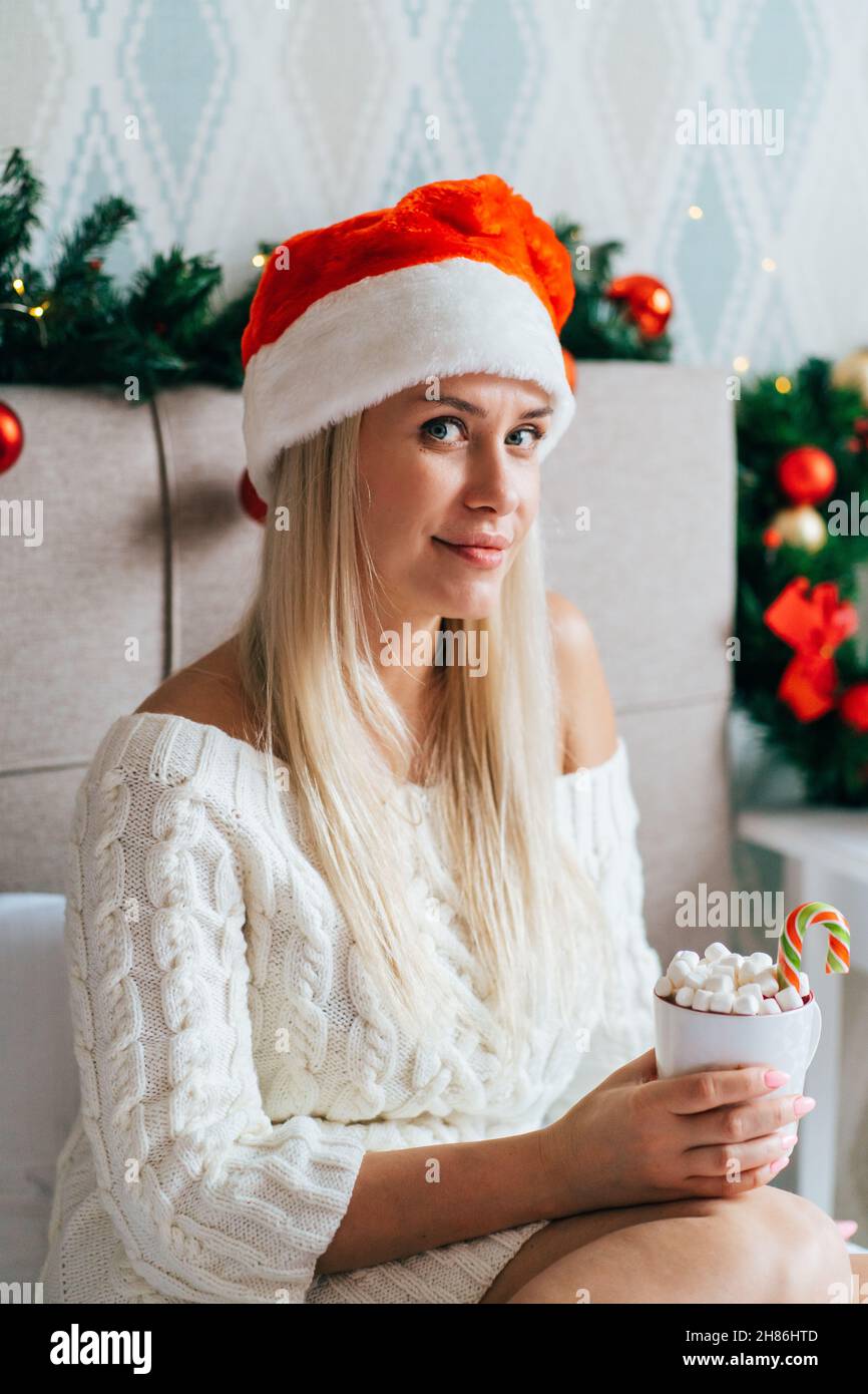 Blonde Frau im weißen Strickkleid Weihnachtsmütze sitzt im weihnachtlich geschmückten Schlafzimmer auf dem Bett und trinkt heißen Kakao mit Marschmalchen Stockfoto