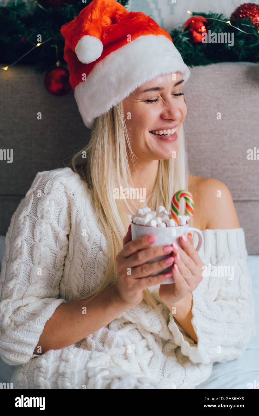 Blonde lächelnde Frau im weißen Strickkleid Weihnachtsmütze sitzt im weihnachtlich geschmückten Schlafzimmer auf dem Bett und trinkt heißen Kakao mit Marschmalchen Stockfoto