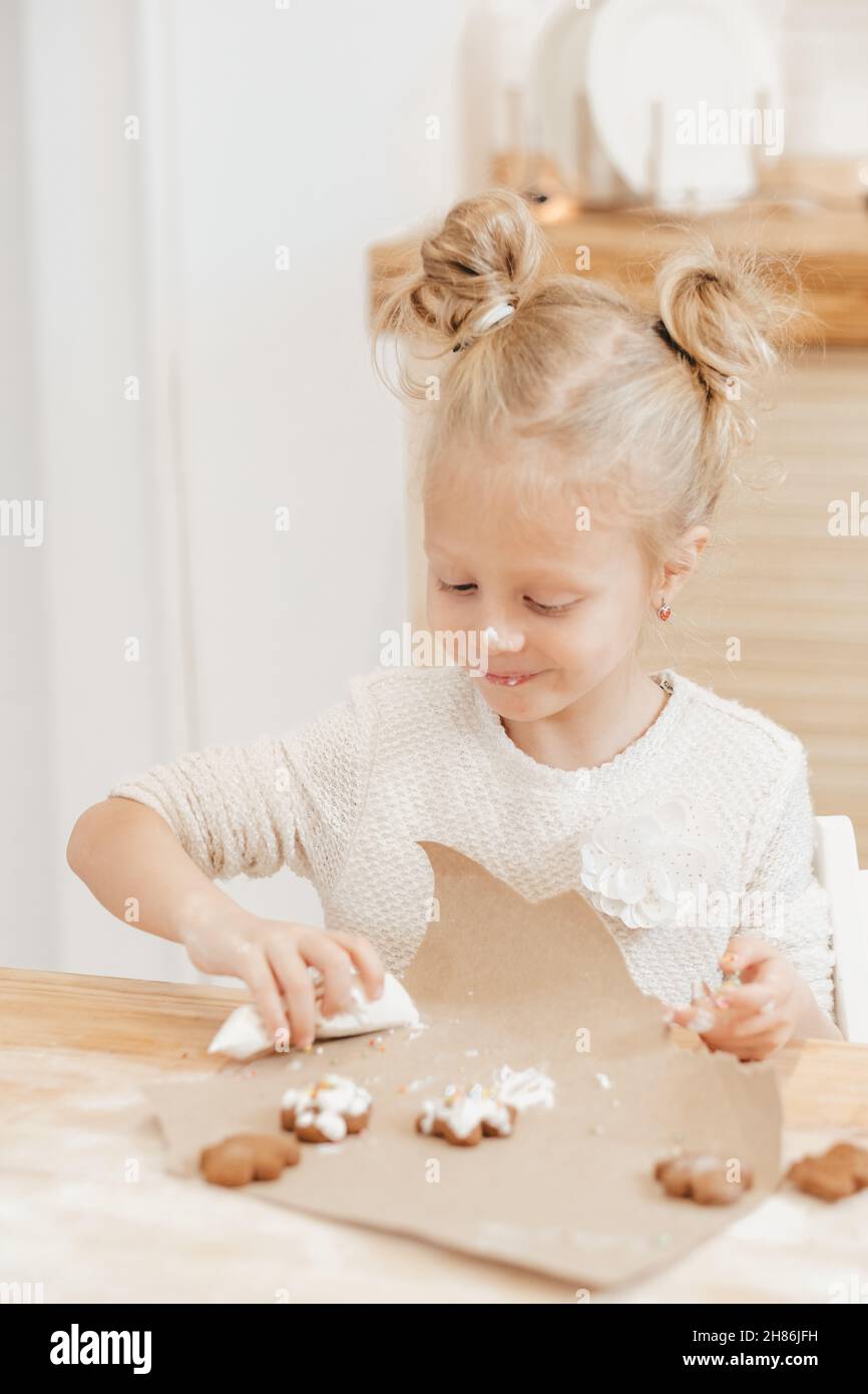 Blonde Mädchen malt Lebkuchen mit weißer Creme. Das Kind schmückt Weihnachtskekse mit Glasur auf einem Holztisch in der Küche. Stockfoto