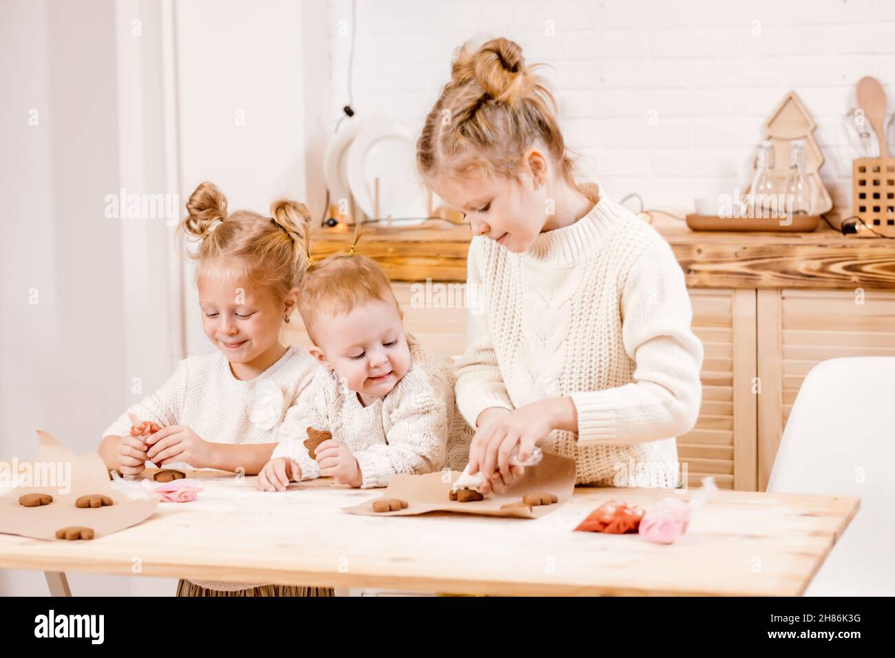 3 blonde Mädchen schmücken Weihnachtskekse mit Glasur auf einem Holztisch in der Küche. Die Freundinnen kochen zusammen in der Küche mit Christus Stockfoto