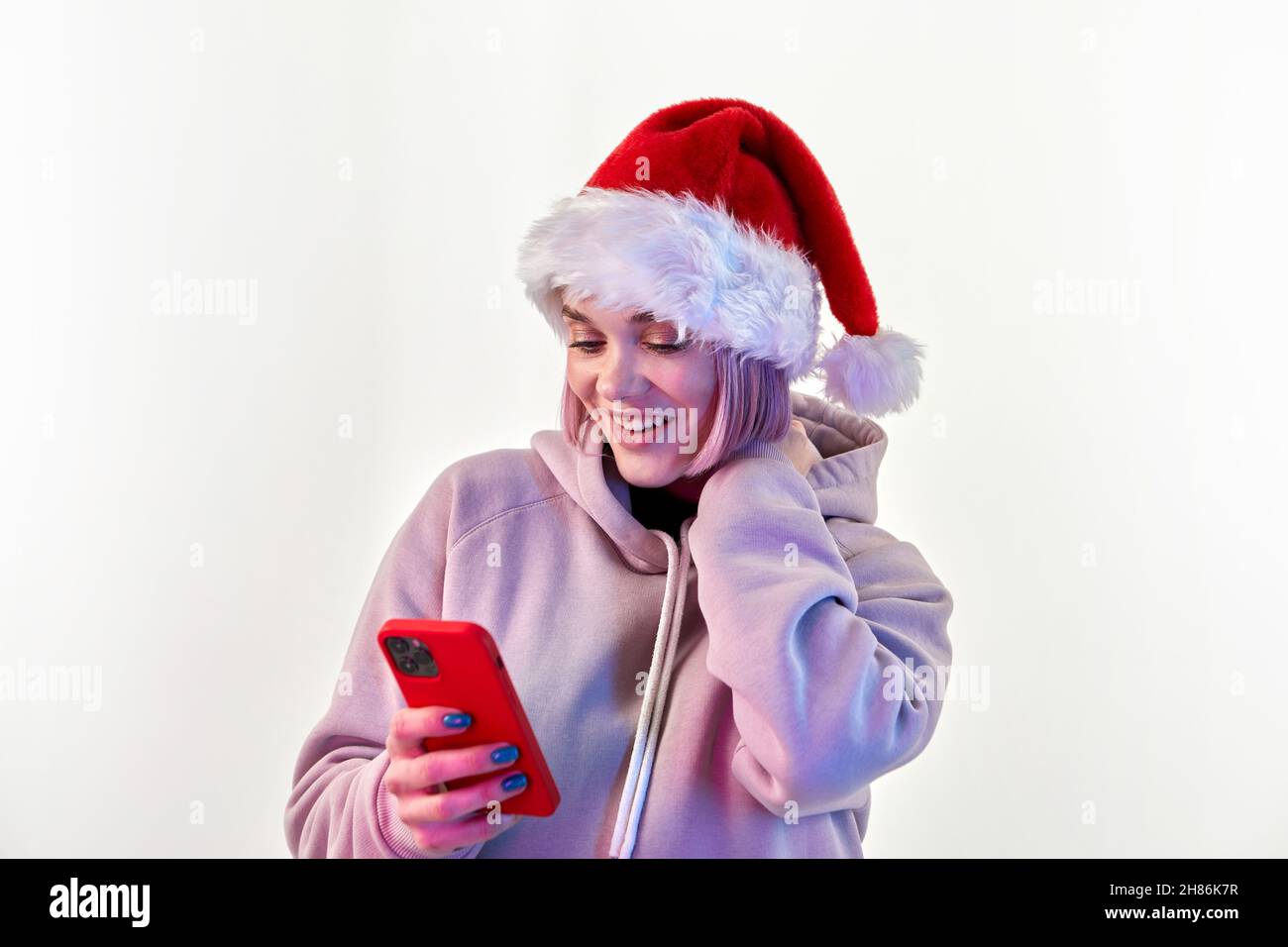 Staunend attraktive junge Frau in weihnachtsmann Hut mit rosa Haar in lila Hoodie mit Handy auf weißem Hintergrund Stockfoto