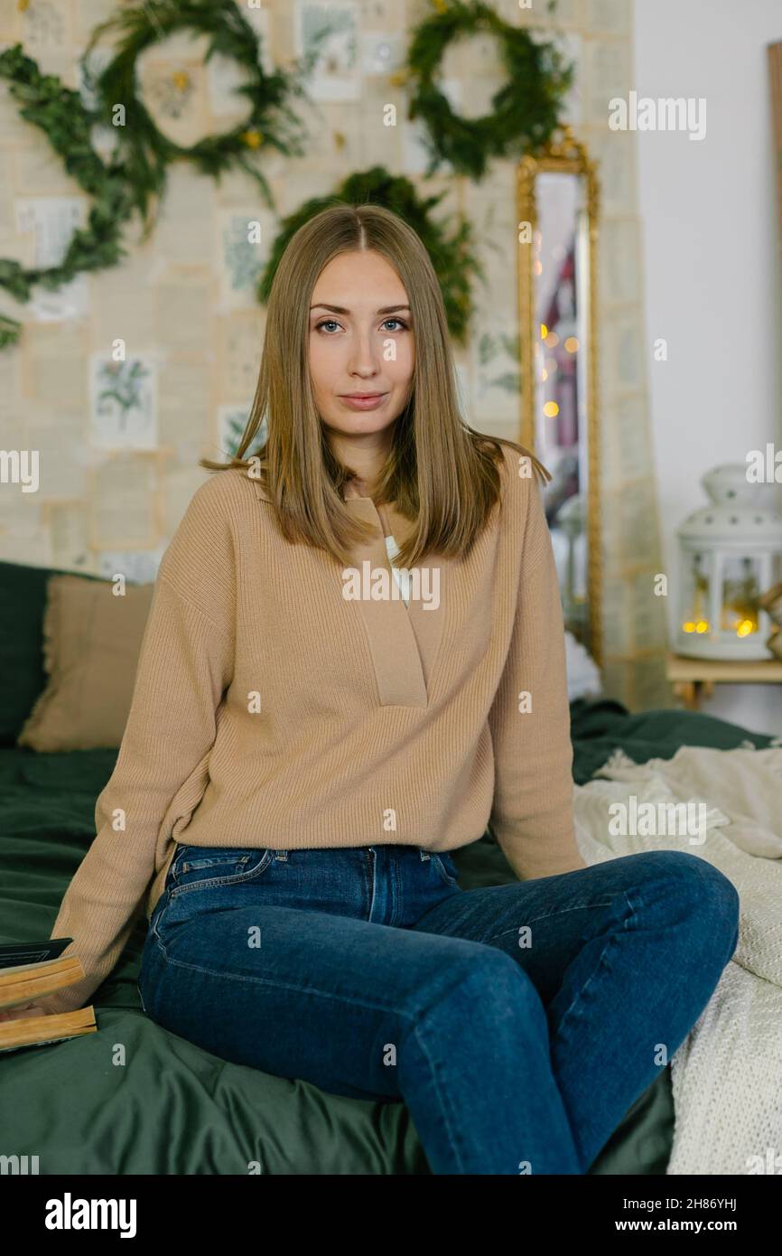 Porträt der jungen schönen kaukasischen Frau auf dem Bett im Dekorationsraum für Weihnachtsferien sitzen. Stockfoto