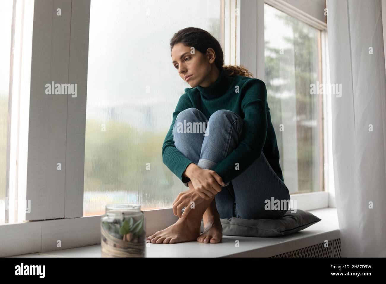 Traurige hispanische Dame sitzt barfuß auf dem Fensterbrett fühlen sich abwesend gesinnt Stockfoto