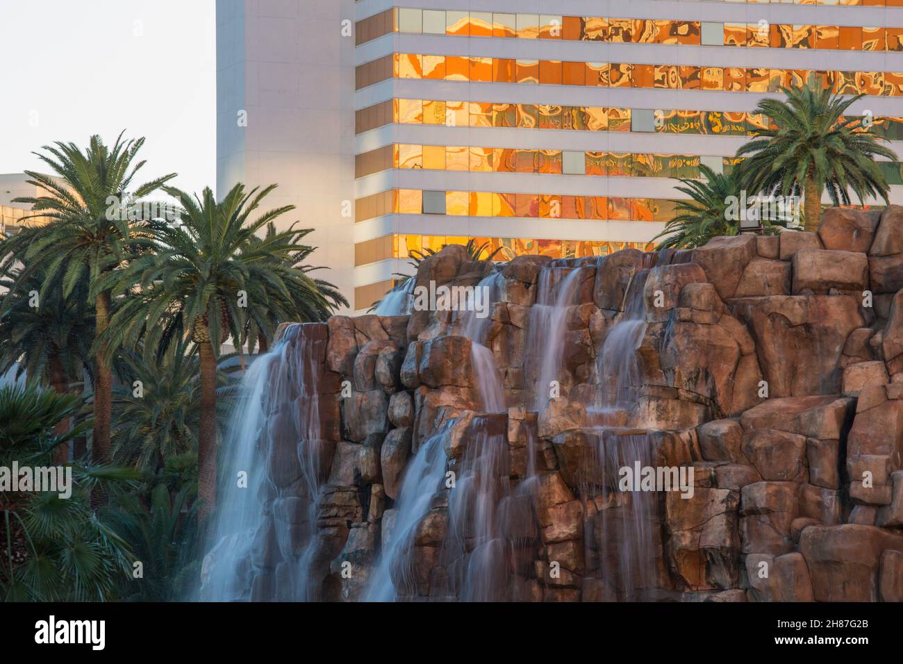 Las Vegas, Nevada, USA. Ornamentaler Wasserfall, der über rote Felsklippen vor dem Mirage Resort und Casino stürzt, Sonnenuntergang, Palmen im Hintergrund. Stockfoto