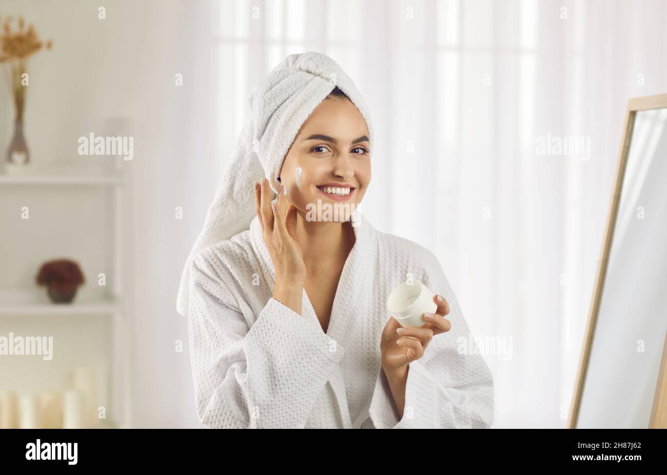 Fröhliche junge Frau mit einem Handtuch auf dem Kopf lächelnd, während sie Anti-Falten-Creme auf ihr Gesicht auftragen Stockfoto