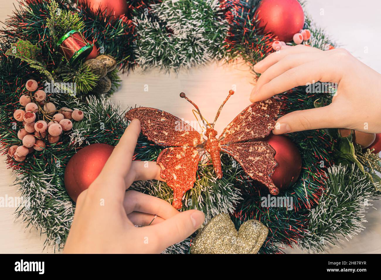 Weihnachtskranz vor der Tür. Draufsicht auf weibliche Hände, die den Weihnachtskranz mit roten Kugeln mit Schmetterlingsform dekorieren. Familienadvent Stockfoto