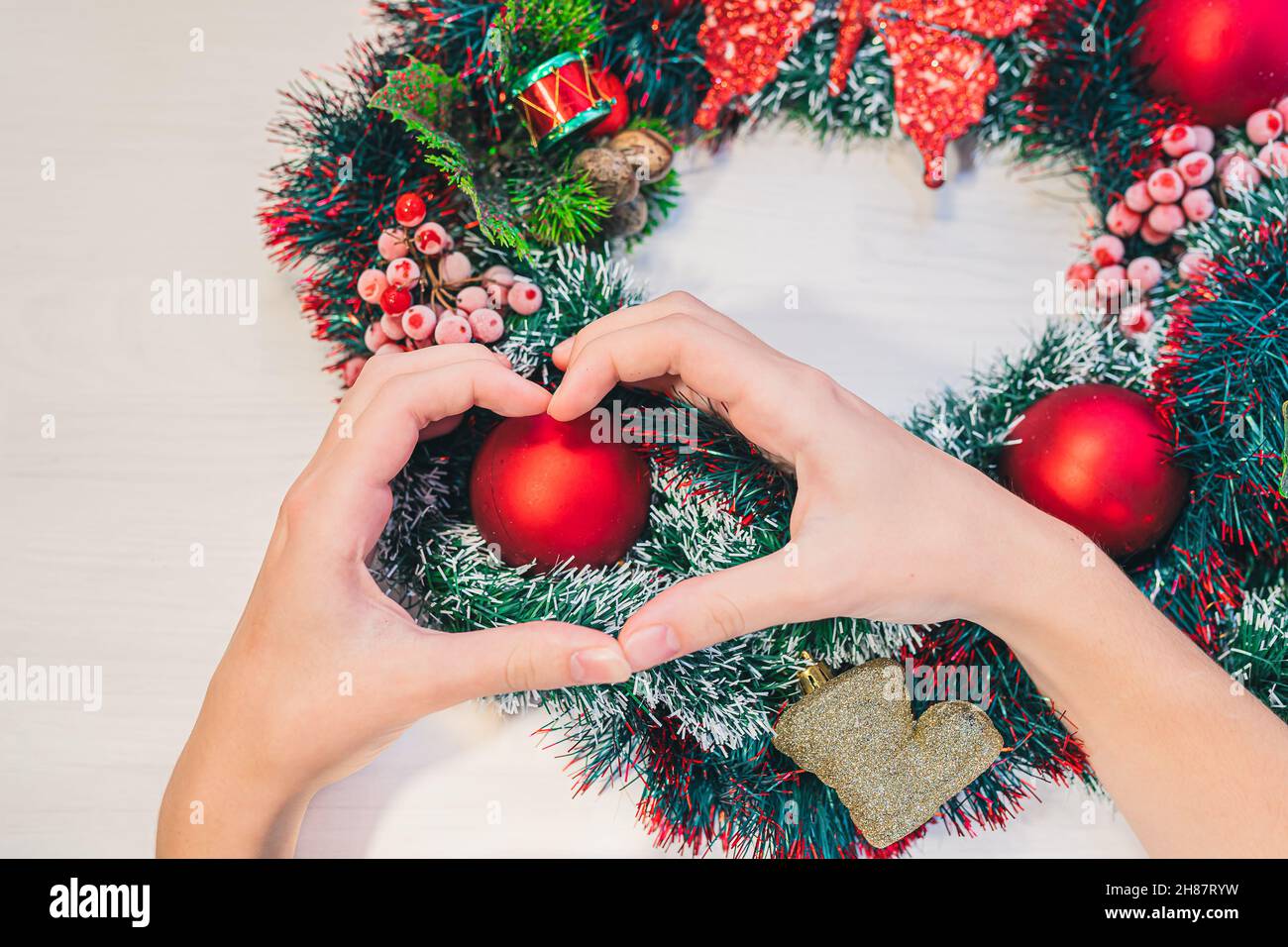 Weihnachtskranz auf Holzhintergrund. Frauenhänden zeigen auf dem Hintergrund des Weihnachtskranzes mit roten Kugeln ein Herzsymbol. Stockfoto