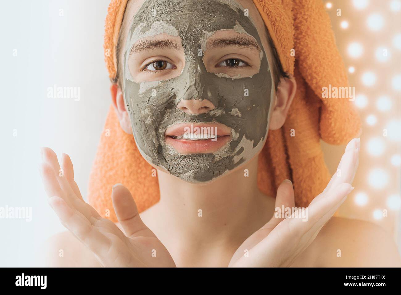 Kaukasisches Model Mädchen Teenager in Handtuch auf dem Kopf trägt kosmetische Maske aus grünem Heilerde auf verschwommenem Hintergrund von Lichtern der festlichen Girlande Stockfoto