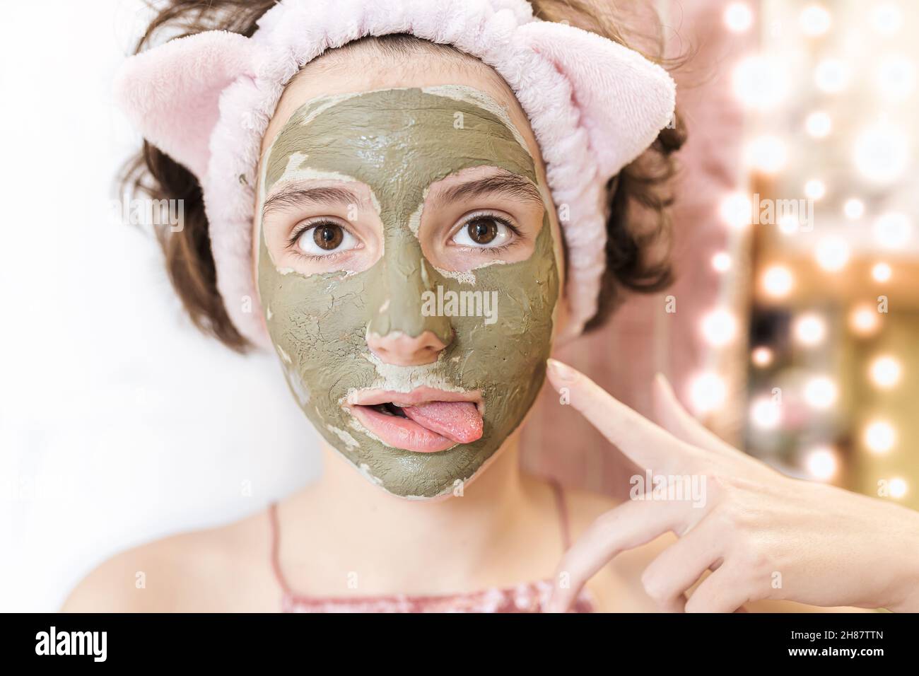 Ein attraktives Teenager-Mädchen in Maske aus grünem Ton macht Gesichter auf verschwommenem Hintergrund von Weihnachtsgirlande. Spa-Behandlungen zu Hause Stockfoto