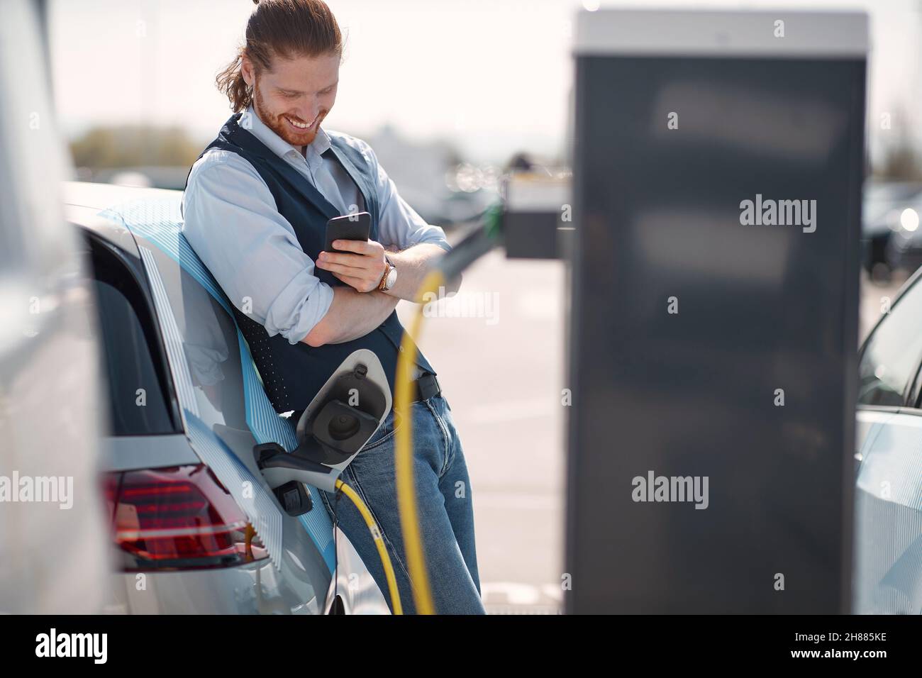 Junger lächelnder Mann mit Smartphone in der Nähe eines Elektroautos an der Ladestation Stockfoto