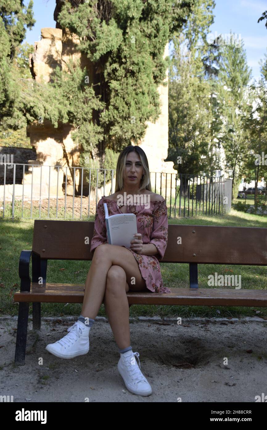 Frau, die während eines sonnigen Tages ein Buch in einem Park liest, schaut auf eine Kamera, schaut auf ein Buch, sitzt auf einer Bank, an einem Winter-, Sommertag Stockfoto