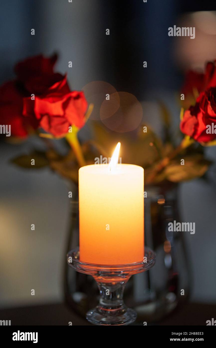 Nahaufnahme der weißen Kerze auf dem Tisch in der Nähe von schönen roten Rosen für ein romantisches Abendessen. Konzept der gemütlichen Atmosphäre mit schönen Kerzen und Blumen Stockfotografie