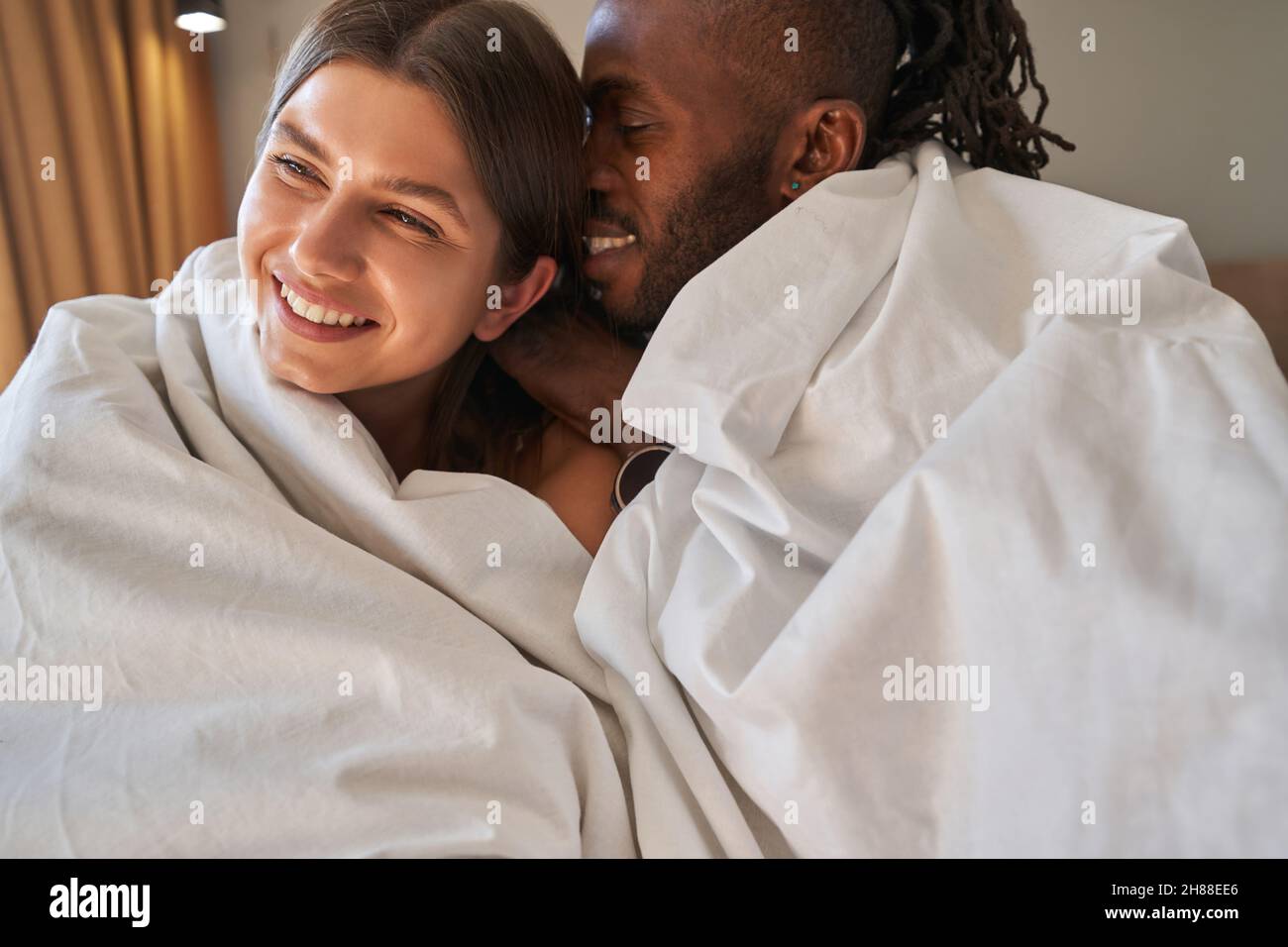 Romantisches Paar mit gemischter Rasse, eingewickelt in Bettdecken, das im Bett sitzt Stockfoto