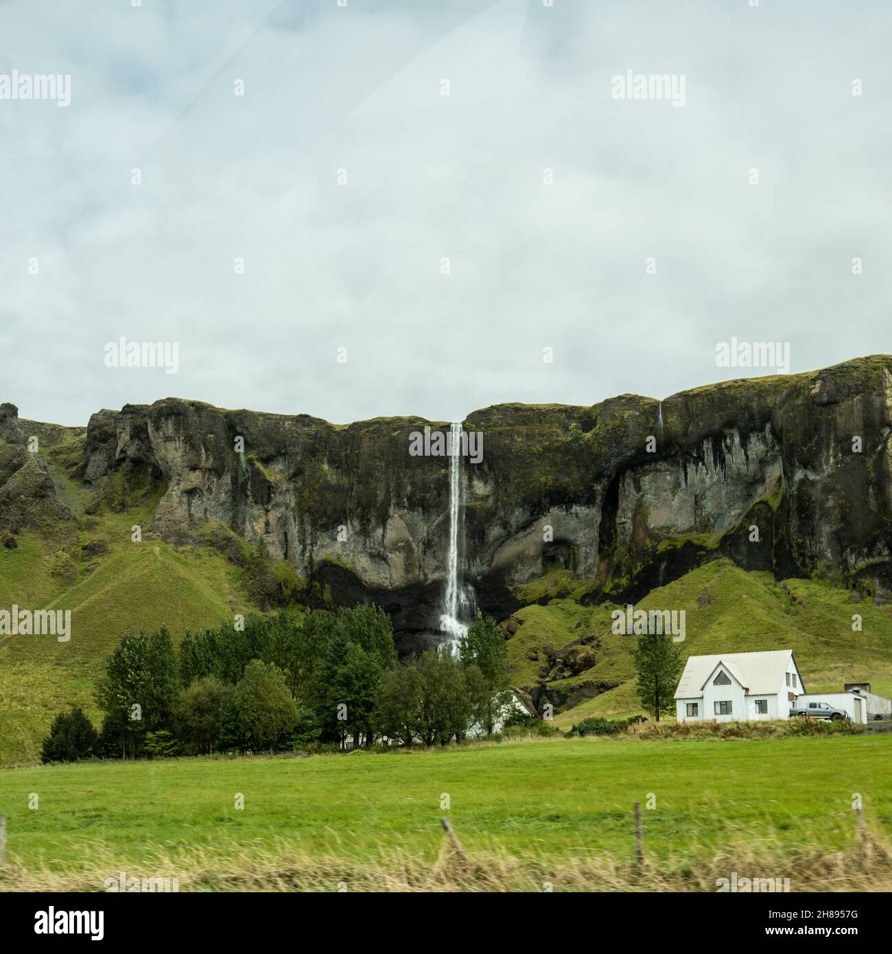 Malerischer Wasserfall stürzt einen Berg hinunter mit einem kleinen weißen Häuschen in der Nähe, Island. Stockfoto
