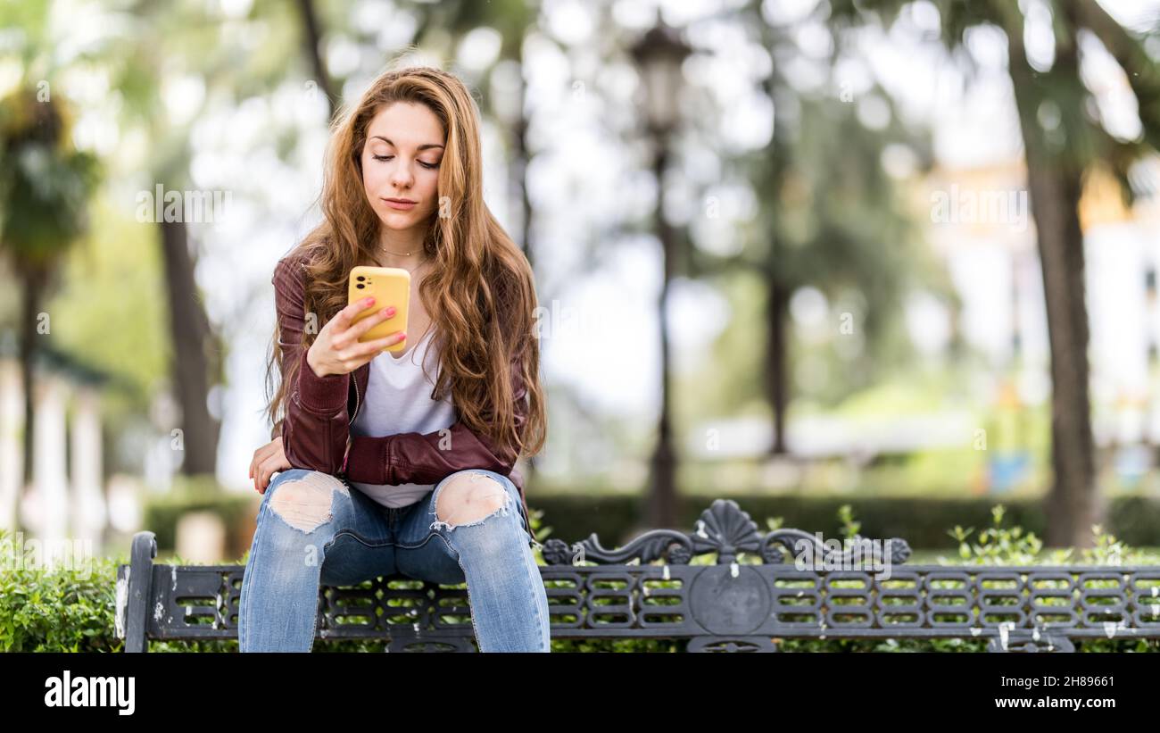 Junges Mädchen, das auf ihr Smartphone schaut, das auf einer Bank im Park-Format mit Kopierplatz sitzt Stockfoto
