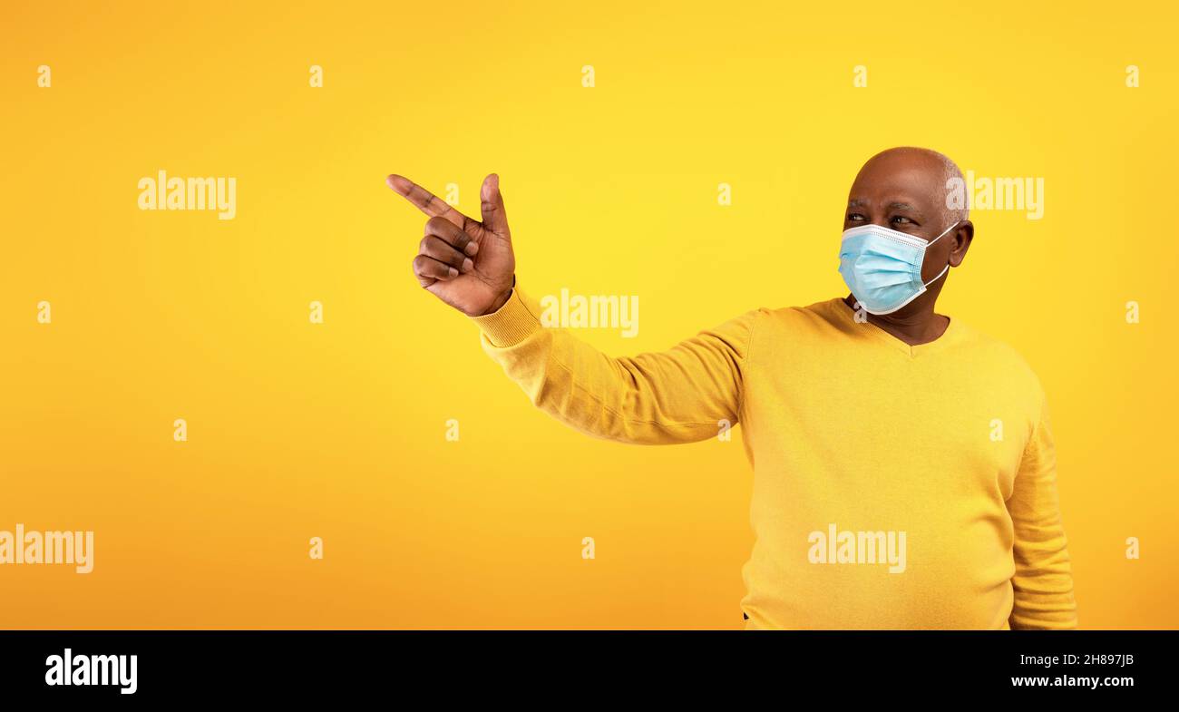 Älterer schwarzer Mann in chirurgischer Gesichtsmaske, der auf leeren Raum für Text oder Werbung über orangefarbenem Studiohintergrund, Banner, zeigt Stockfoto