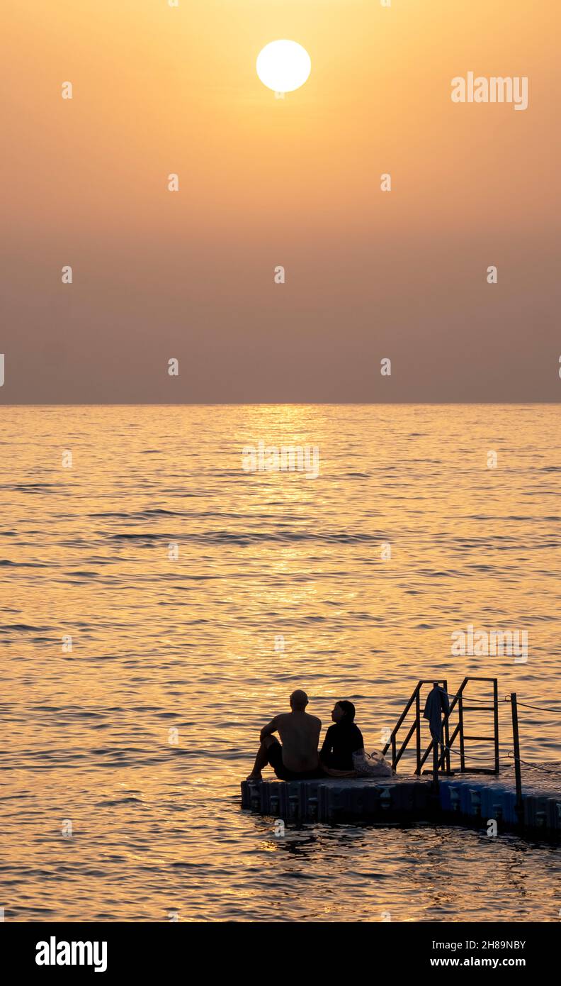 Ein Paar beobachtet den Sonnenuntergang, während sie zusammen auf einem Ponton in Paphos, Zypern, sitzen. Stockfoto