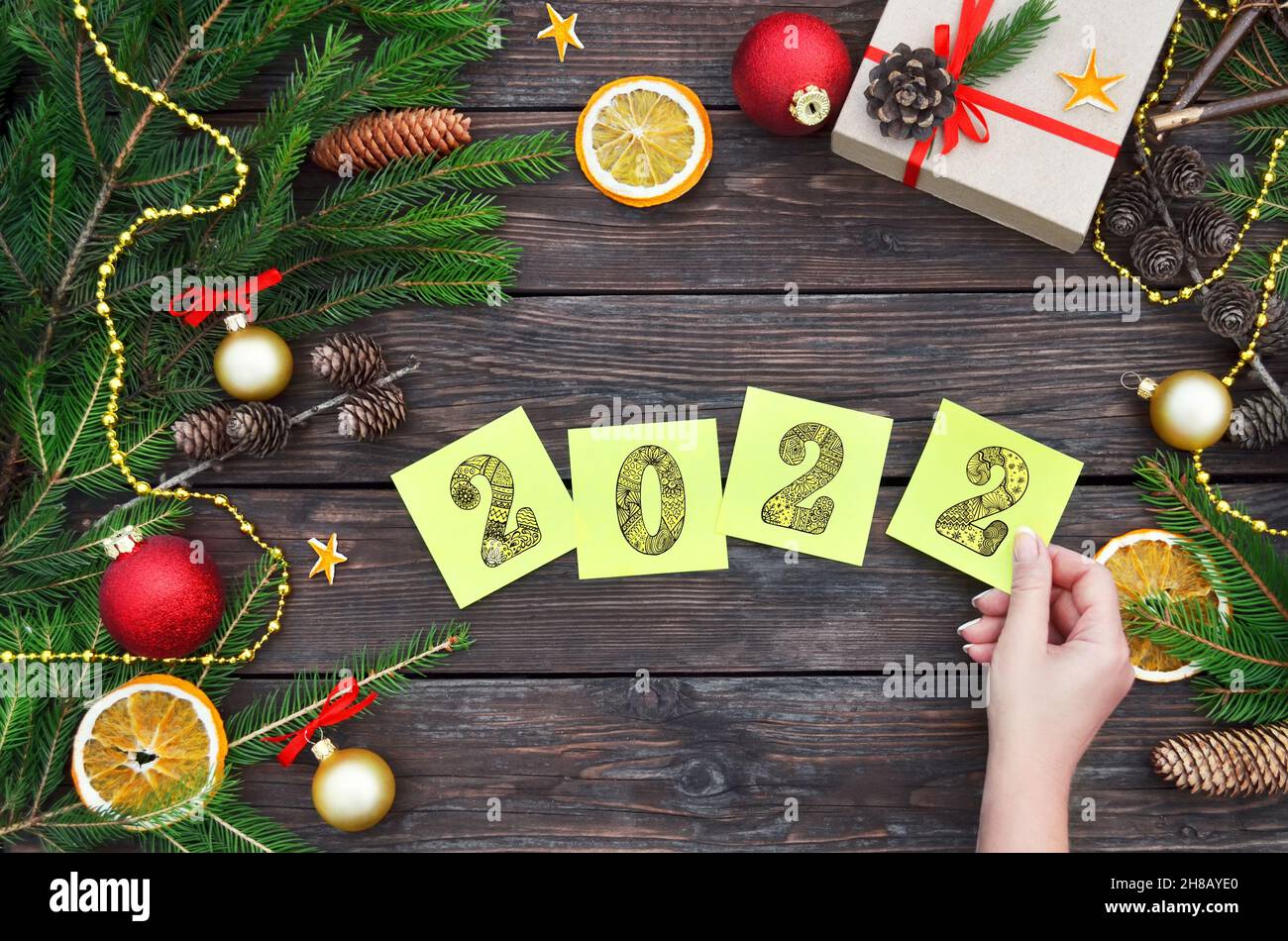 Eine Frauenhand legt gelbe Haftnotizen mit den Zahlen 2022 auf einen festlichen Holzhintergrund. Neujahrskomposition mit Geschenkbox, Tannenzweigen, Zapfen Stockfoto