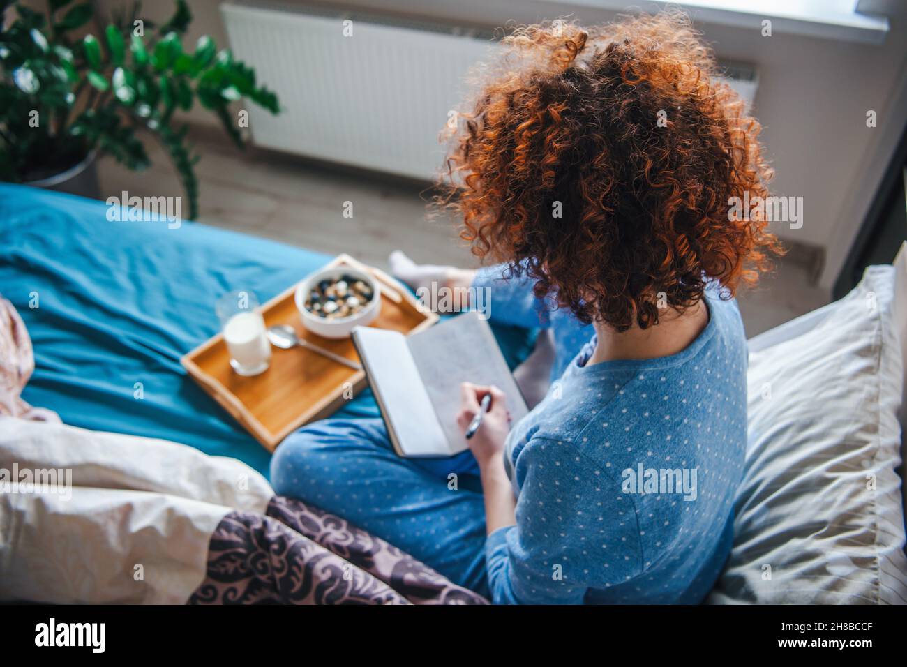 Rückansicht einer lockigen Frau, die auf dem Bett sitzt und etwas in seinem Notizbuch notiert. Tablett mit leckerem Frühstück auf dem Bett. Stockfoto