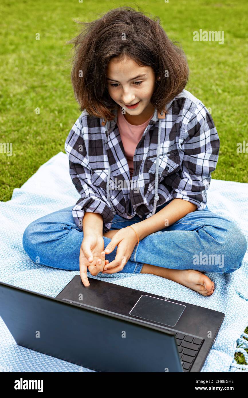 Ein süßes Teenager-Mädchen, das auf dem Gras sitzt und an einem Laptop arbeitet. Ein Schulmädchen schaut sich einen Internetkurs an und studiert aus der Ferne. Das Mädchen nutzt Wireless Stockfoto