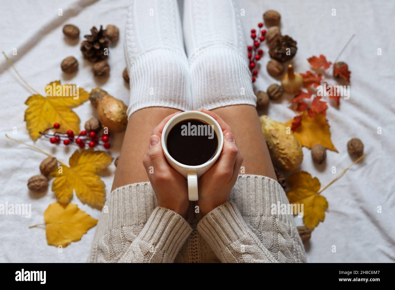 Frau Hände halten eine Tasse Kaffee auf den Beinen in wollenen Wintersocken in einem gemütlichen Bett mit Herbstblättern zu Hause dekoriert. Nahaufnahme, selektiver Fokus Stockfoto
