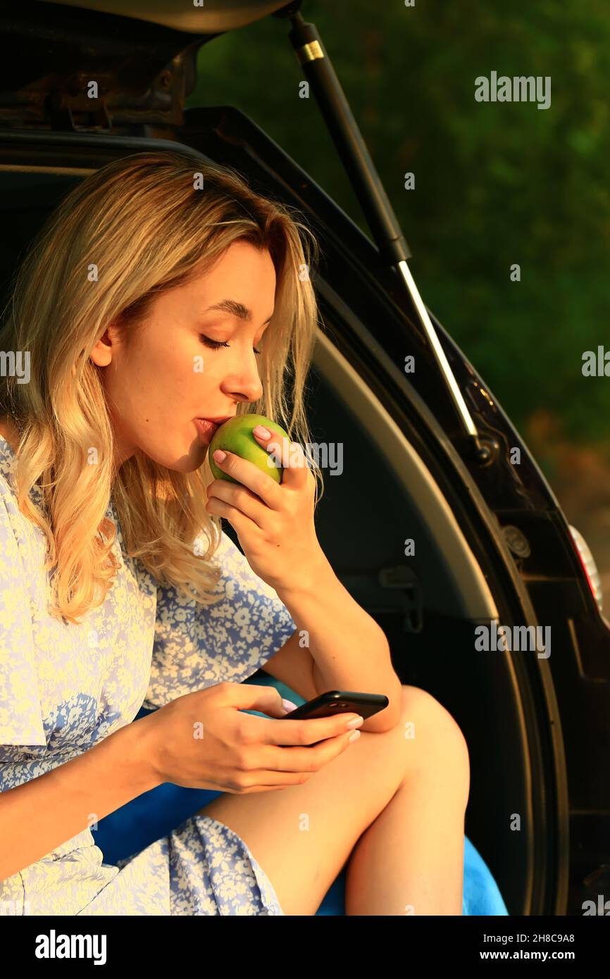 Blondine in einem blauen Kleid hat eine Pause im offenen Kofferraum eines Autos und einen Bissen Apfel sitzen. Eine Reise allein in der Nähe des Hauses auf dem Land. Stockfoto