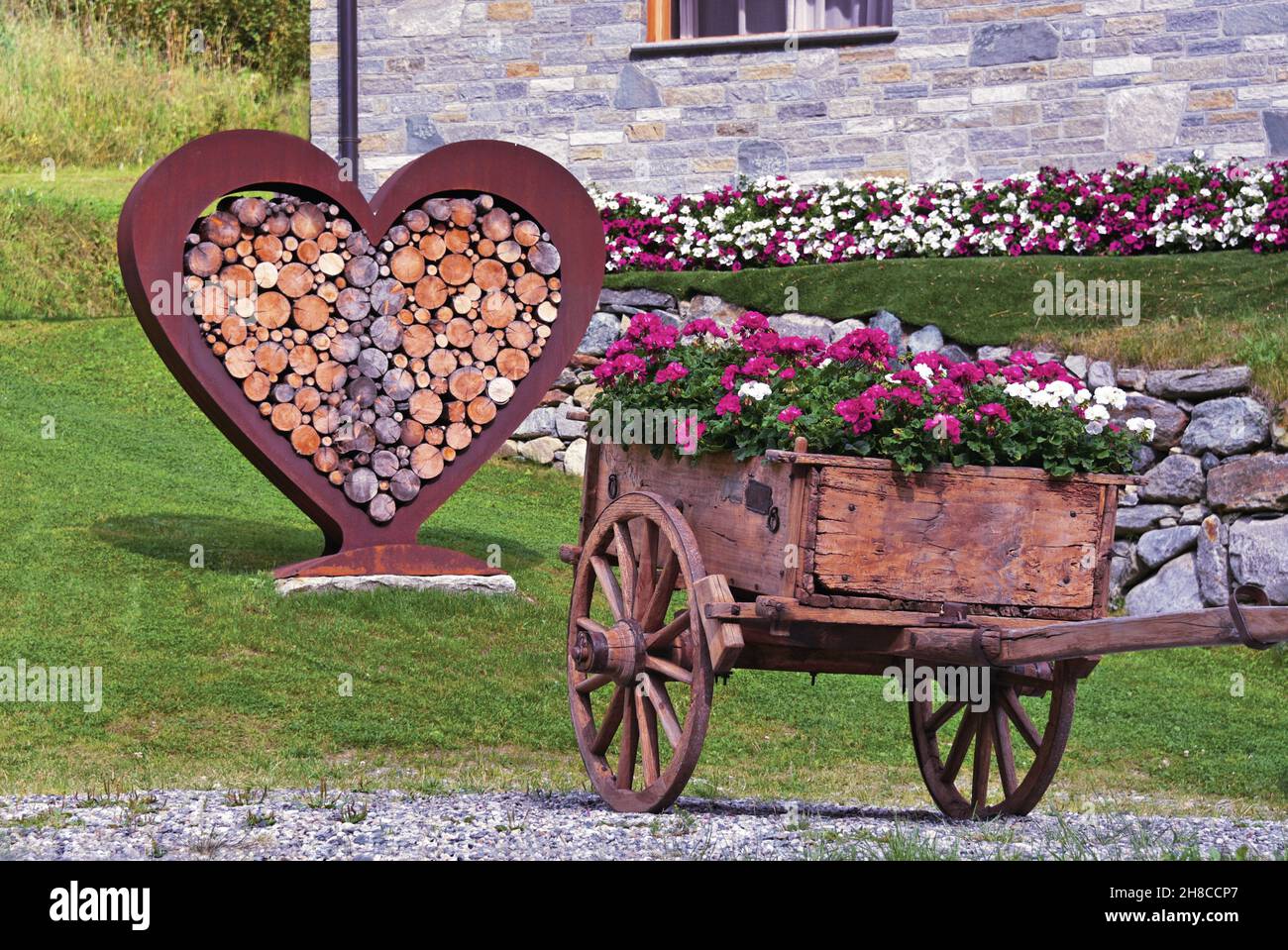 Gepflanzt Holzkarren und Metall Herz gefüllt mit Brennholz als Dekoration in einem Garten, Italien, Aostatal, Gressoney Stockfoto