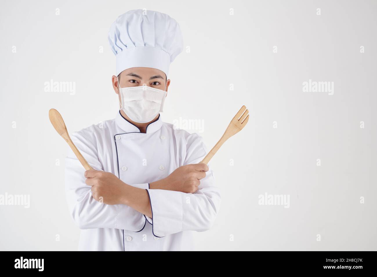 Portrait des fröhlichen Küchenchefs in medizinischer Maske mit Holzlöffel und Spachtel Stockfoto