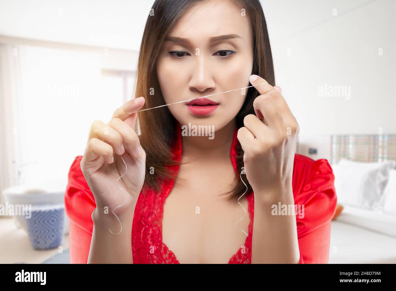 Schönes Mädchen mit Zahnseide entfernt Zahnbelag. Eine Frau in einem roten Seidengewand, die Zähne mit Zahnseide auf einem Schlafzimmerhintergrund putzt. Stockfoto