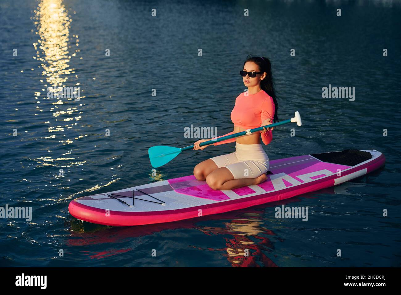 Junge kaukasische Frau in Sportkleidung, die lange Paddel zum Schwimmen auf dem Boot auf dem Stadtsee verwendet. Reflexion vom Sommeruntergang auf der Wasseroberfläche. Workout im Freien. Stockfoto
