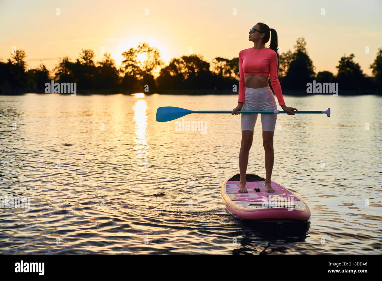 In voller Länge Porträt einer aktiven Frau in Sportkleidung, die während des atemberaubenden Sonnenuntergangs auf dem Boot mit Paddel schwebt. Junge Brünette mit sportlichen Aktivitäten auf dem Wasser am Abend. Stockfoto