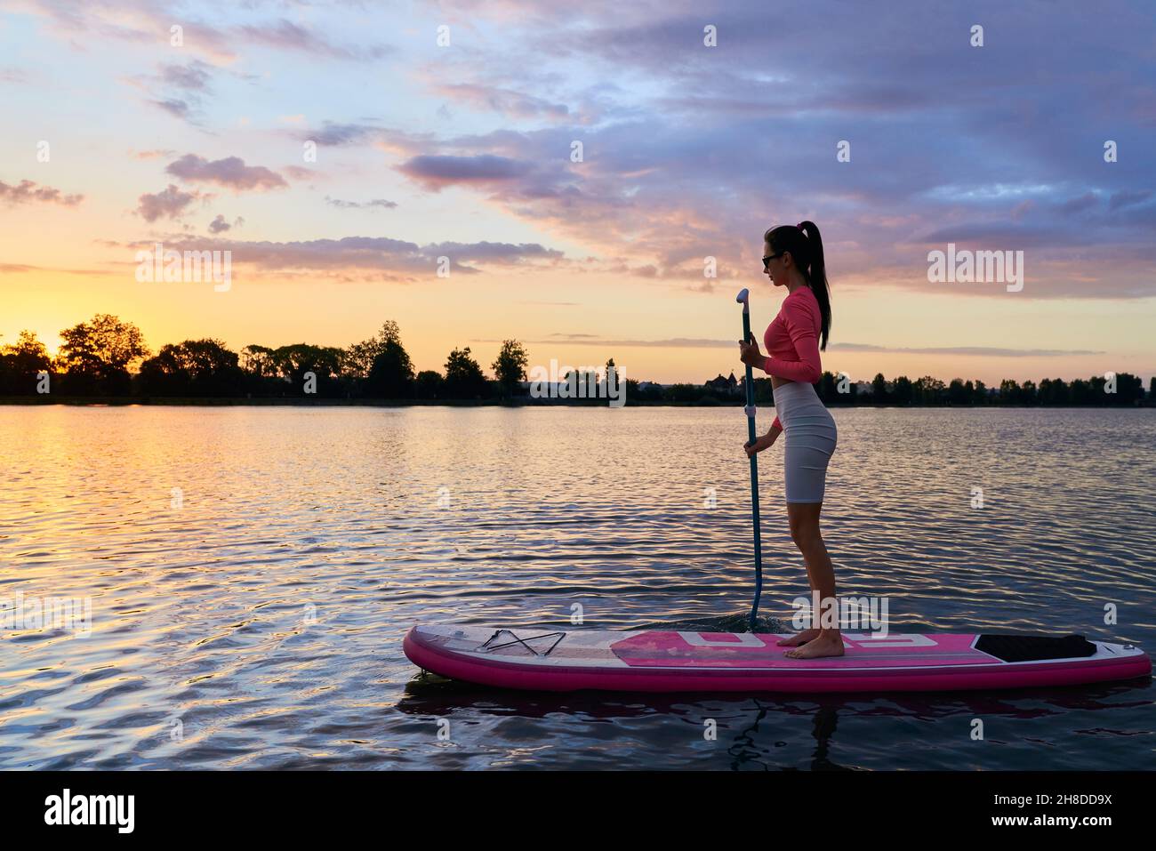 Aktive junge Frau mit braunem Haar, die im Schwimmen auf dem Brett mit Paddel und Sommeruntergang auf dem Hintergrund trainiert. Konzept von Menschen, Sport und gesunder Lebensweise. Stockfoto