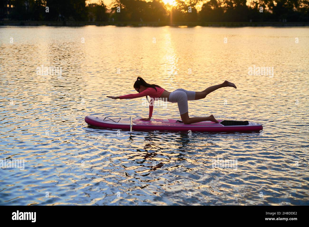 Fit junge Frau in Sportkleidung tun Yoga-Übungen auf bunten Brett mit erstaunlichen Sonnenuntergang über dem See. Konzept von Mensch, Balance und Energie. Stockfoto